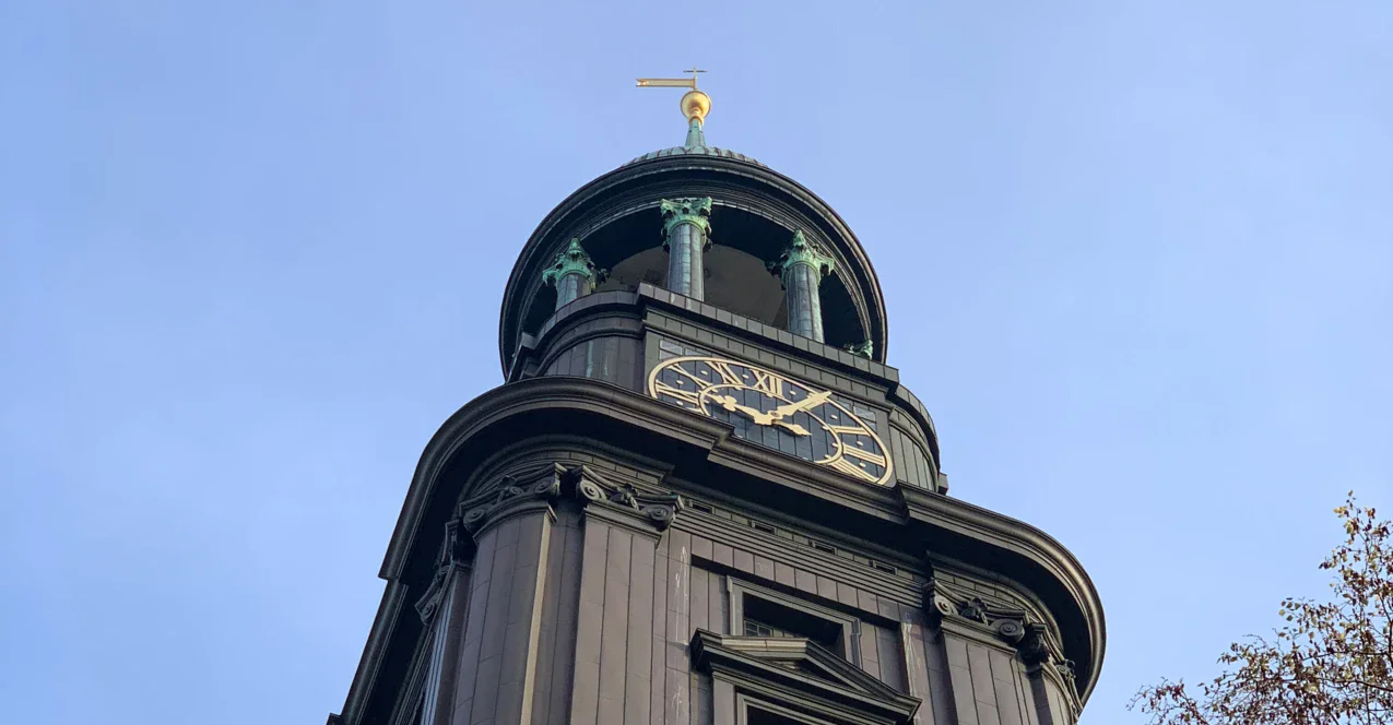 Eine Aufnahme der Spitze des Kirchturms vom Michel. Dieser ragt in dunklem Grau mit einer großen goldenen Uhr in den blauen Himmel empor. 