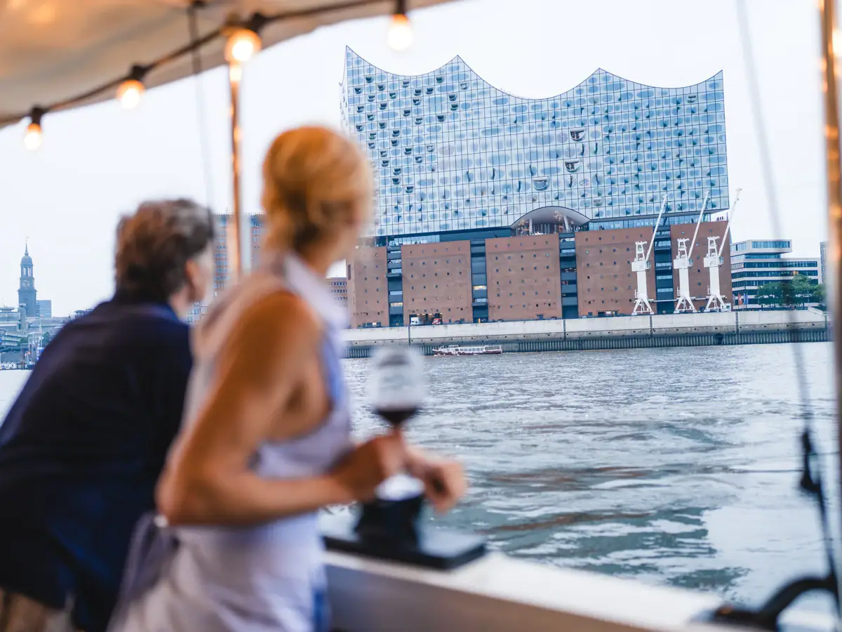 Ein Mann und eine Frau stehen an der Reling der Barkasse MS Jacob und blicken auf die Elbphilharmonie und die Elbe.