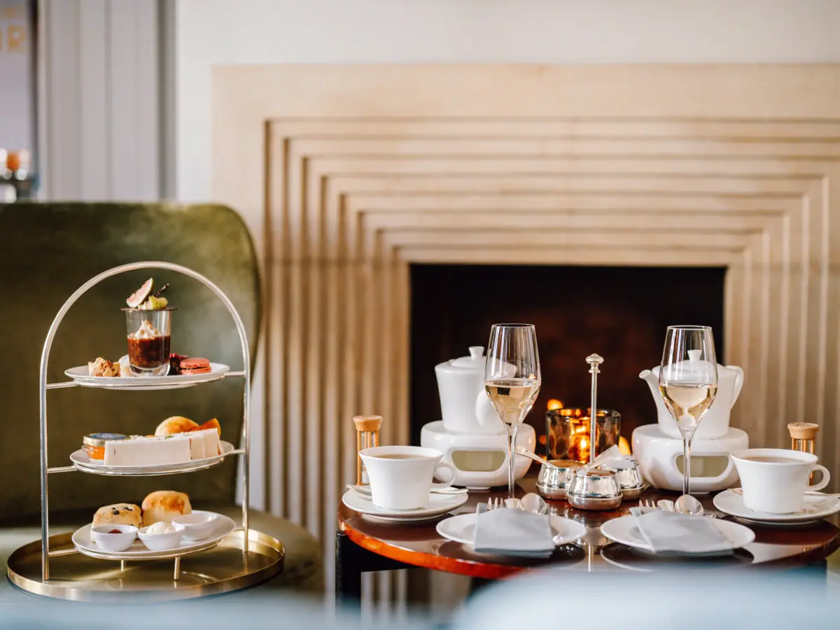 Afternoon Tea Wohnhalle Teezeit im Hotel Louis C. Jacob. Vor dem Kamin in der Wohnhalle steht auf einem Tisch eine Etagere mit herzhaften und süßen Köstlichkeiten, Tee und zwei Gläsern mit Champagner.