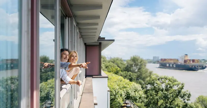 A man and a woman look out of a window of the Elbe Suite at the Louis C. Jacob onto the Elbe.