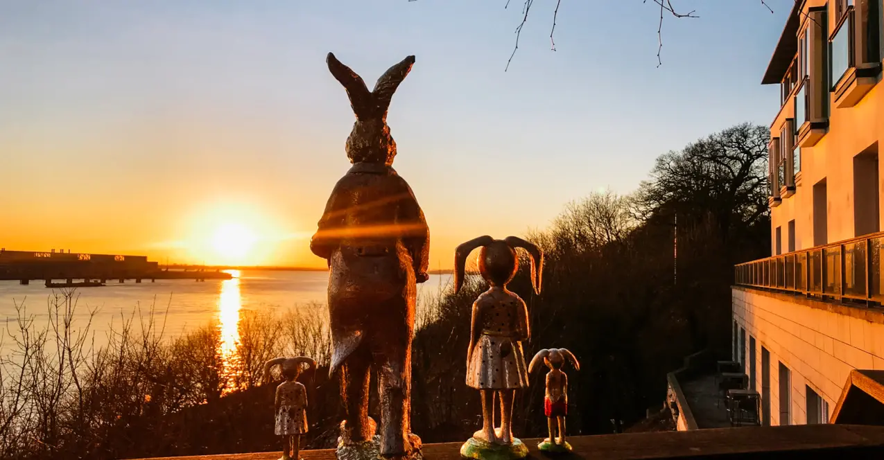 Statue eines Hasen und zwei Kinder auf einem Geländer, im Freien mit Bäumen und Wasser im Hintergrund.