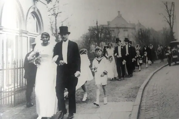 Heinrich Louis Jacob Eine Gruppe von Menschen, darunter eine Braut im Hochzeitskleid und ein Mann im Anzug, geht auf einem Bürgersteig.