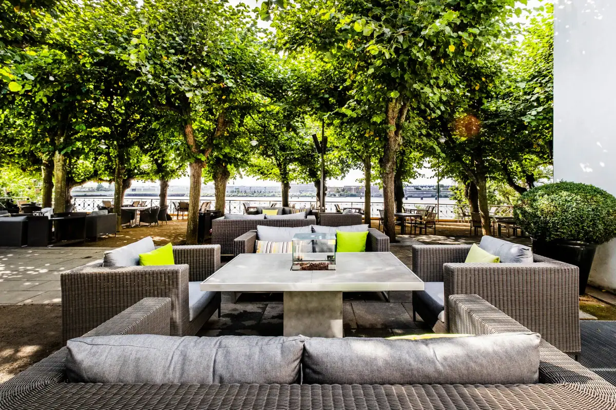 Tische und Sofas stehen unter freiem Himmel auf der Lindenterrasse mit grünen Linden im Hintergrund.