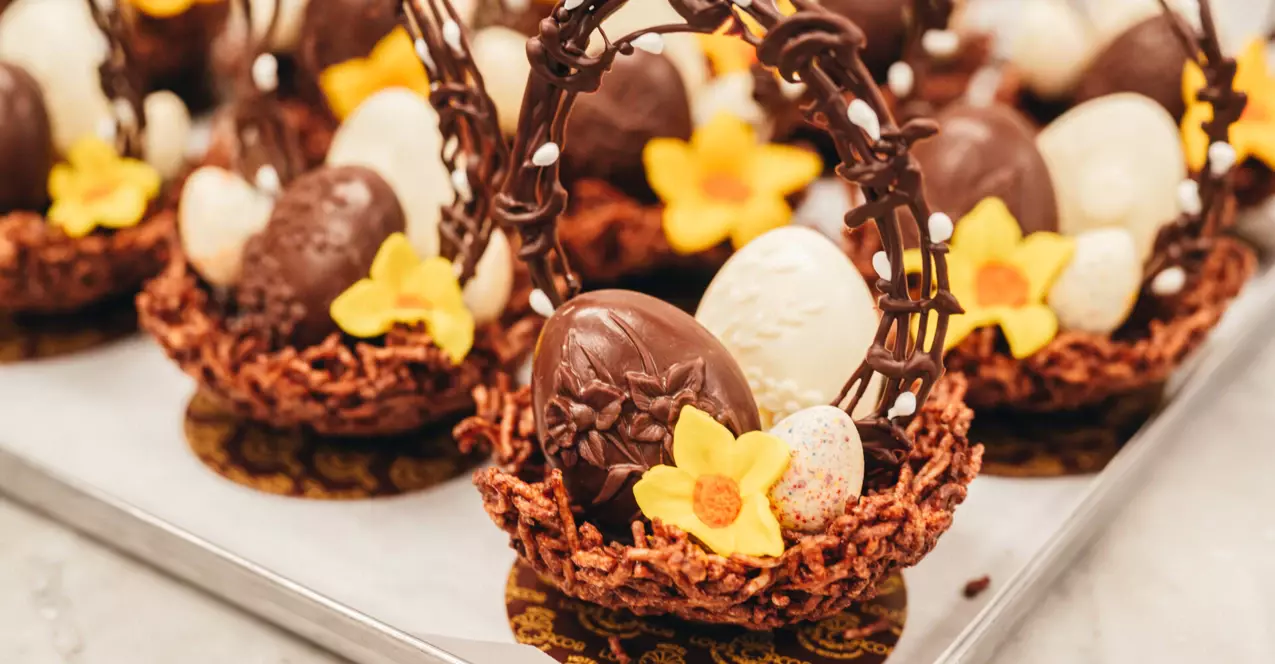 Auf einem Tablett stehen mehrere kunstvoll angerichtete Osterdesserts in Form von kleinen Körbchen mit Schokoladeneiern und einer gelben Blume sowie einem Schokoladenhenkel in form von Ästen mit kleinen Knospen. 