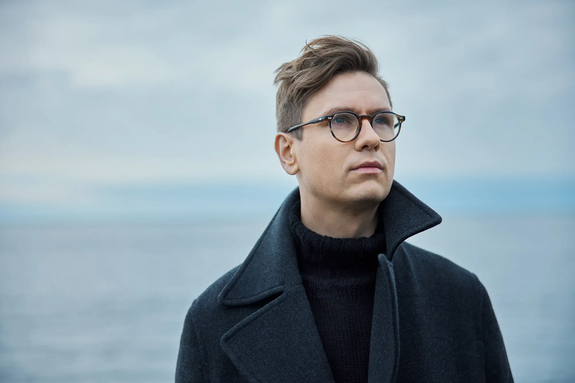 Ein Mann in Mantel und mit Brille steht am Wasser und schaut in die Ferne. 