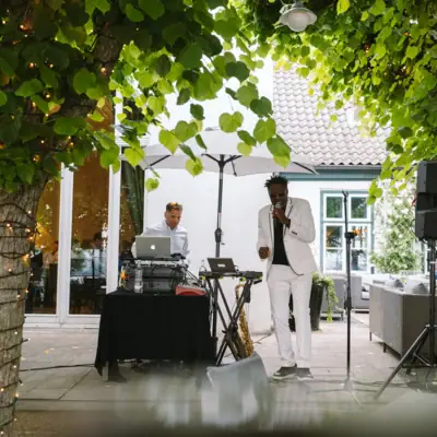 Ein Mann im weißen Anzug singt in ein Mikrofon unter einem Baum und neben ihm steht ein DJ.