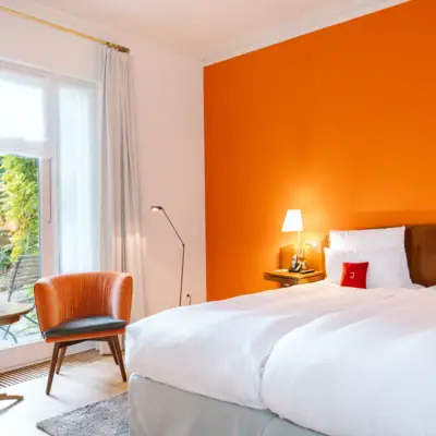 Ein helles Hotelzimmer im Hotel Louis C. Jacob mit gemütlichem Doppelbett, einer orangefarbenen Wand und einer kleinen Terrasse. 