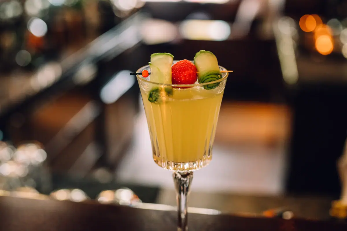 Ein Cocktailglas mit einer gelben Flüssigkeit des Cocktails Mrs. Blankenese mit einer Garnitur von Gurke und Himbeere.