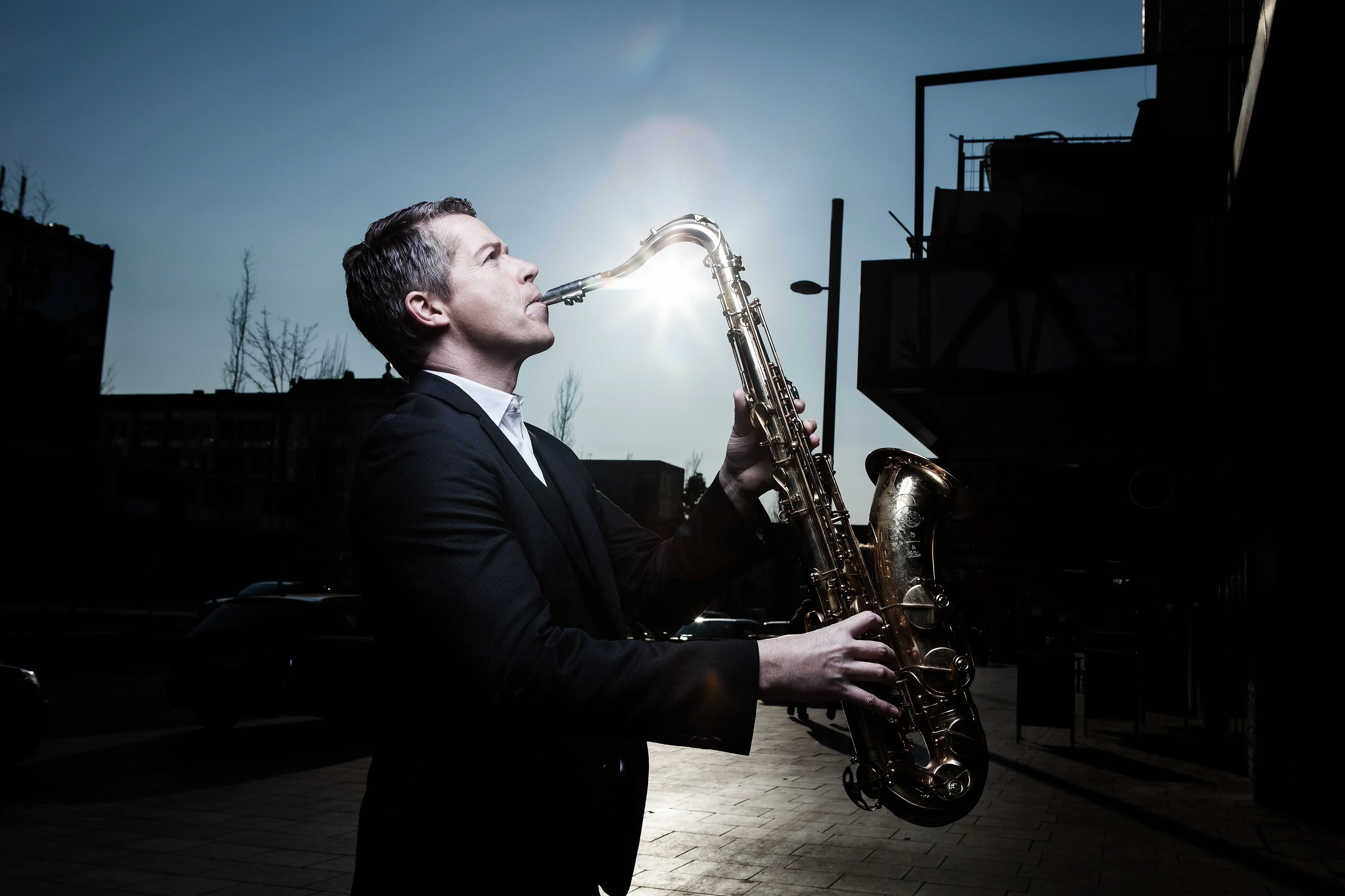 Ein Mann in einem schwarzen Anzug spielt Saxophon und hinter ihm scheint die Sonne.
