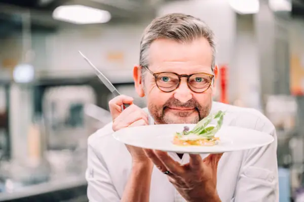 Chefkoch Thomas Martin hält in der einen Hand eine Pinzette und in der anderen hand einen Teller mit einem Gericht.