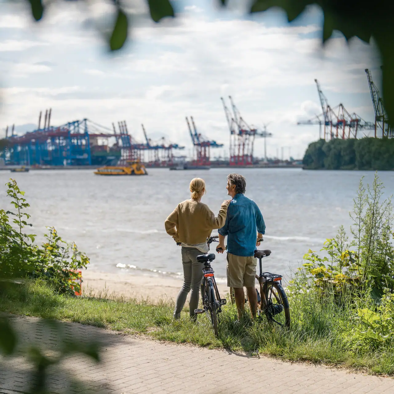 Ein Mann und eine Frau stehen neben Fahrrädern an der Elbe und blicken auf den Hafen von Hamburg.