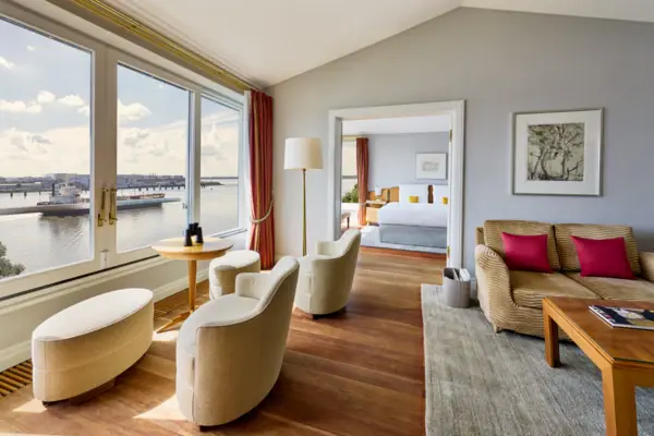 Ein großes Hotelzimmer mit großen Fenster und tollem Blick auf die Elbe sowie mit einem gemütlichen Wohnbereich im Vordergrund und einem separaten Schlafzimmer im Hintergrund.