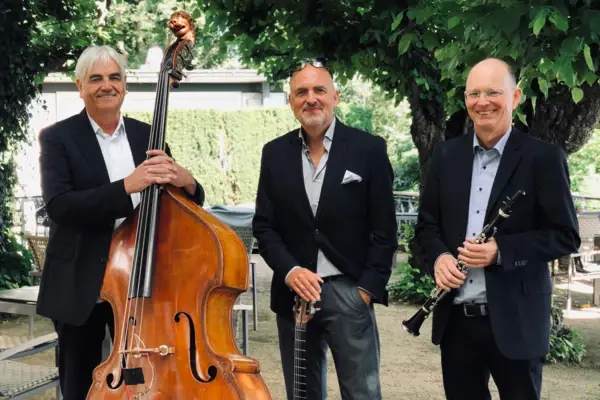 Hamburg String Brothers Drei ältere Männer halten jeweils ein Instrument, wie ein Cello, eine Gitarre und eine Oboe und lächeln in die Kamera.