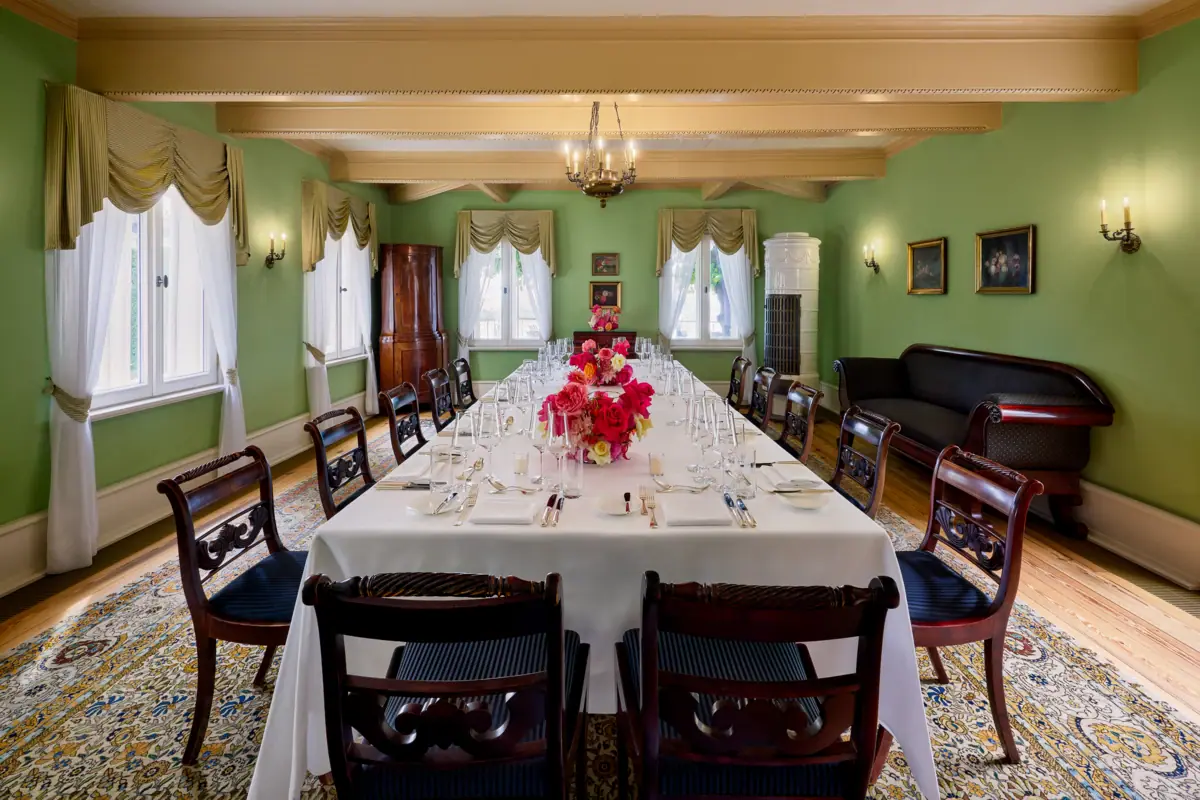 Ein festlicher Veranstaltungsraum mit einer langen Tafel, dunklen Stühlen und roten Blumen auf den Tischen. 