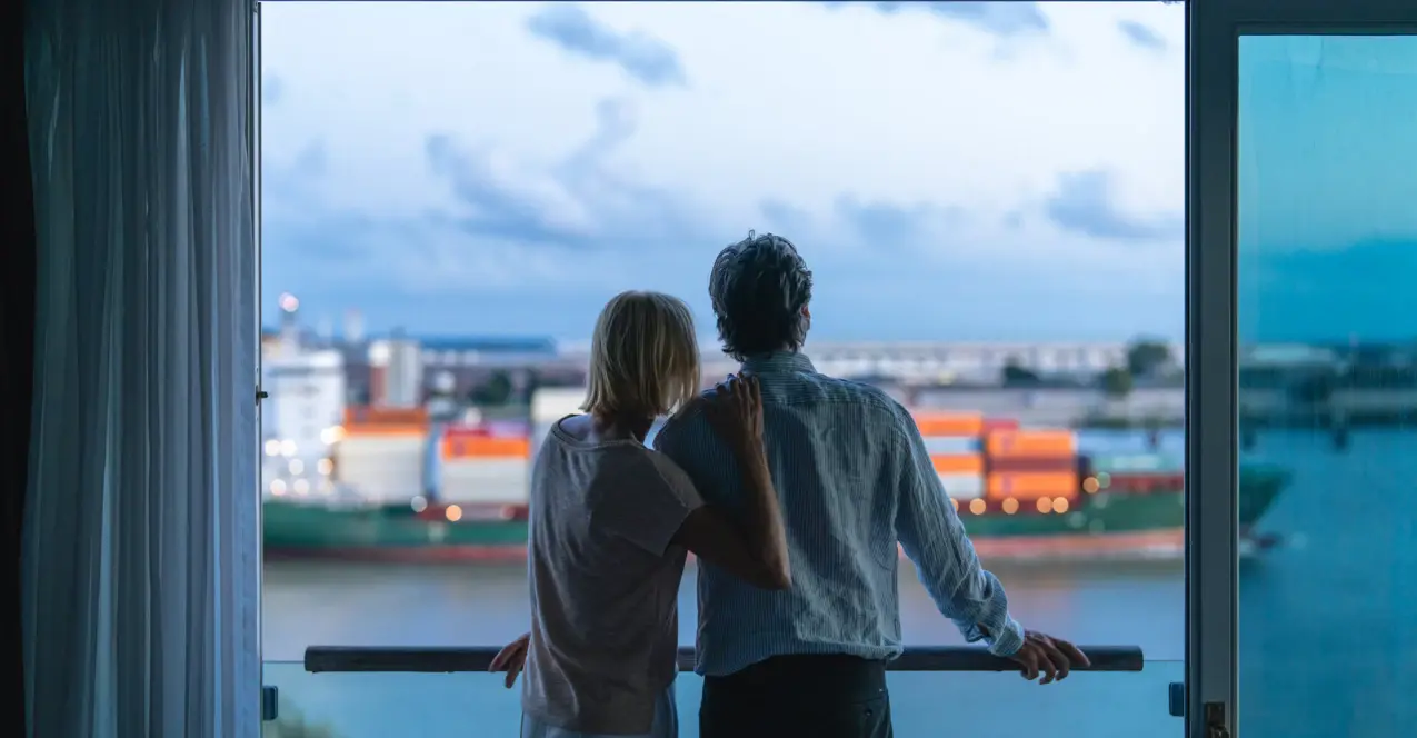 Ein Paar steht aneinander gelehnt an einem offenen Fenster und schaut auf die Elbe, auf der ein Containerschiff vorbei fahrt. 