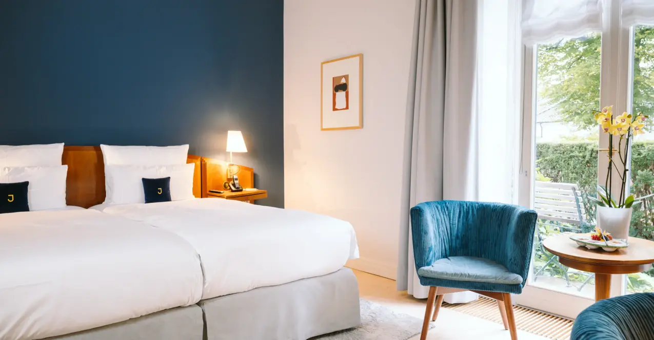 Helles Hotelzimmer mit zwei Betten, dunkelblauer Akzentwand, zwei blauen Sesseln, einem kleinen runden Holztisch mit Orchidee, Nachttischlampen und großem Fenster mit Blick auf einen Garten.