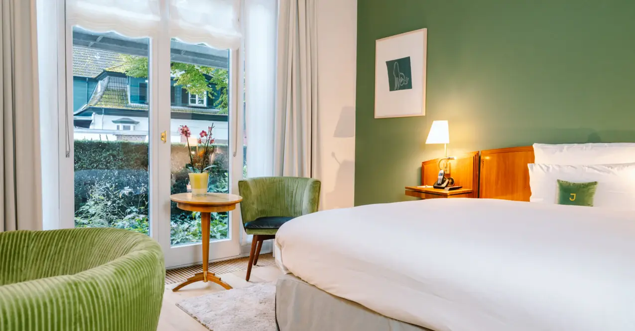 Stilvoll eingerichtetes Hotelzimmer mit grün akzentuierter Wand, gemütlichem Doppelbett, eleganten Sesseln und Blick ins Grüne durch bodentiefe Fenster.