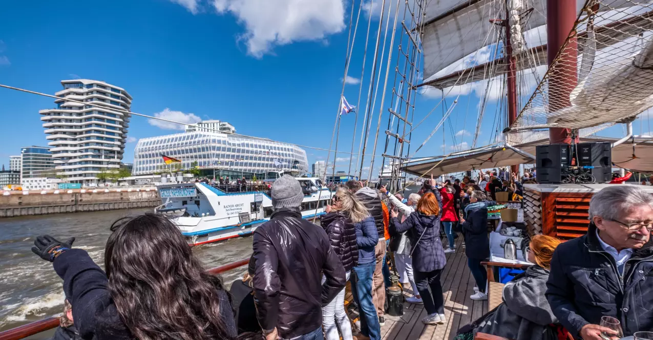 Viele Menschen stehen an Deck eines Schiffes, welches auf der Elbe durch den Hamburger Hafen fährt. Die Passagiere lachen und genießen den Sonnenschein, während ihnen der Fahrtwind durch die Haare weht. 