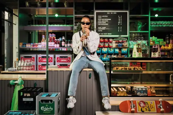 Store & Stuff Eine junge Frau mit Sonnenbrille sitzt auf einer Theke in einem Kiosk nd trinkt aus einem Getränk mit Strohhalm.