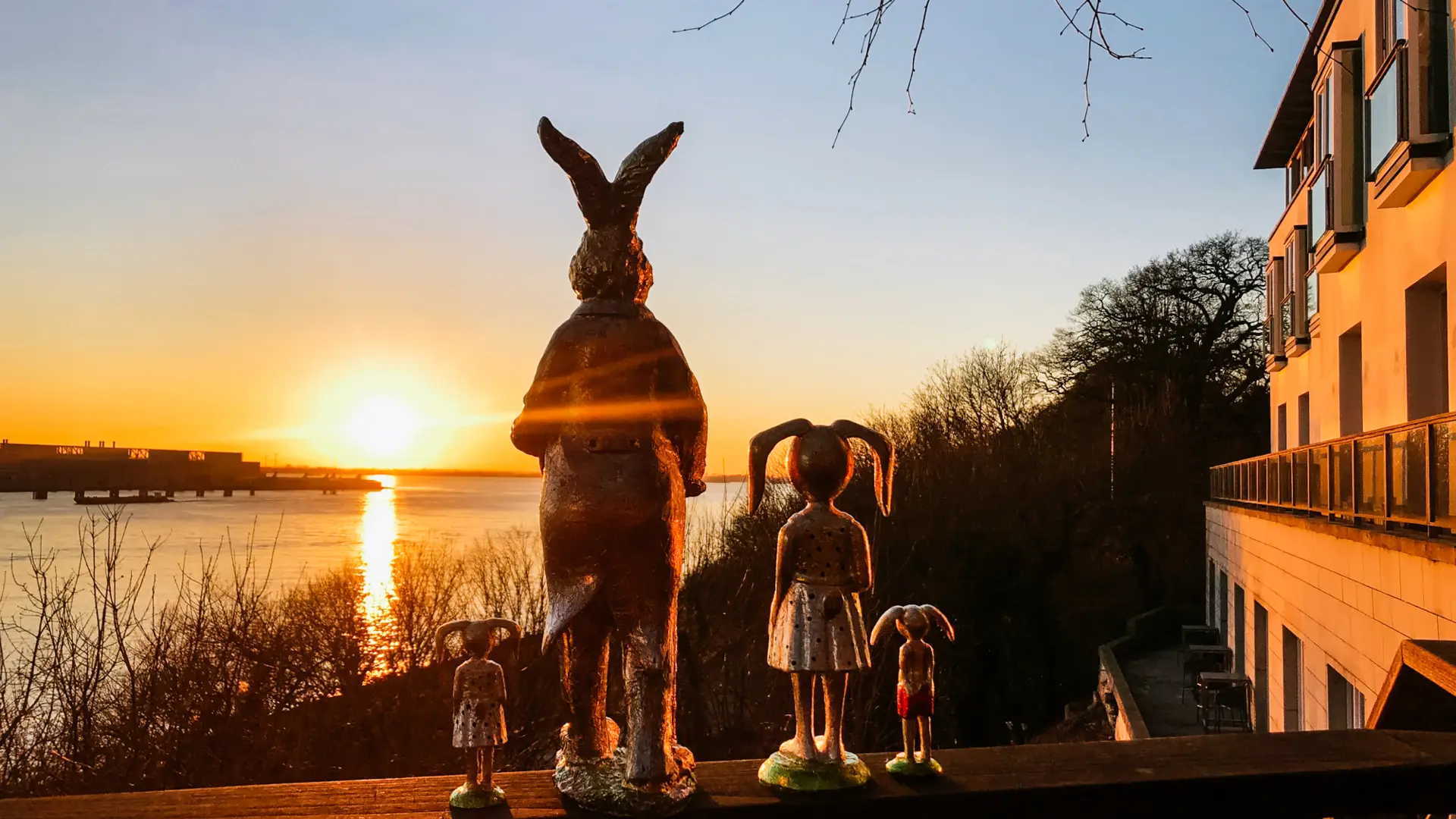 Eine Gruppe von Osterhasenfiguren steht auf dem Geländer einer Terrasse eines Hauses am Wasser. Die Sonne geht auf und taucht das Bild sowie das Wasser in warme goldene Farben.