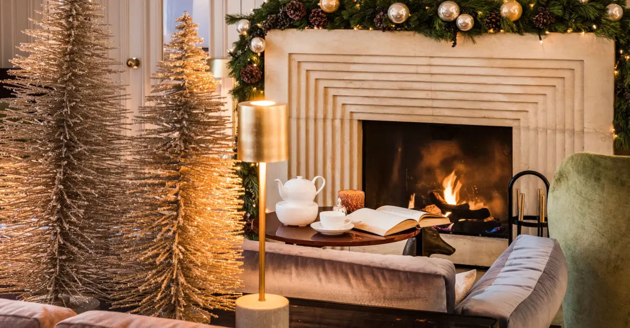 Ein sehr gemütlicher Wohnbereich mit einem brennenden Kamin mit Tannen und Christbaumkugelschmuck. Davor steht ein Sofa mit einem kleinen Holztisch, auf dem ein Buch aufgeschlagen liegt sowie eine Tasse und Teekanne stehen. Kleine goldenen Schmucktannenbäume stehen dekorativ neben einer goldenen Lampe, die warmes Licht spendet.  