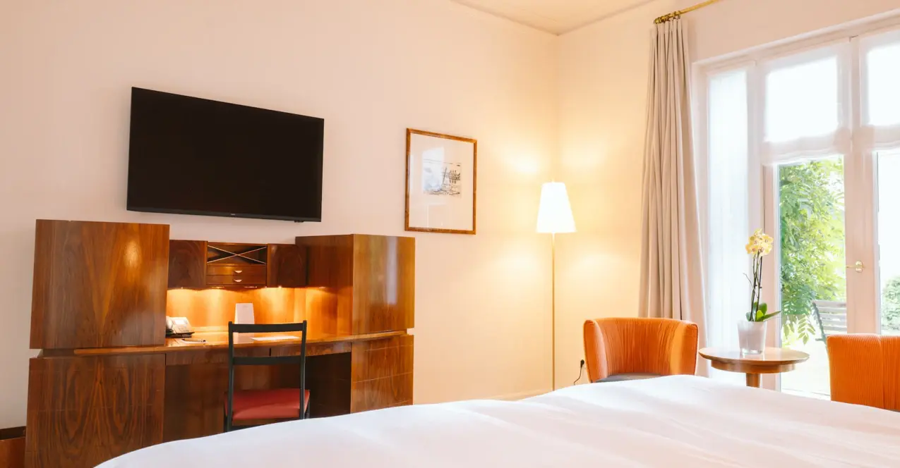 Interior view of a hotel room with large windows, a bed, a desk and a television.