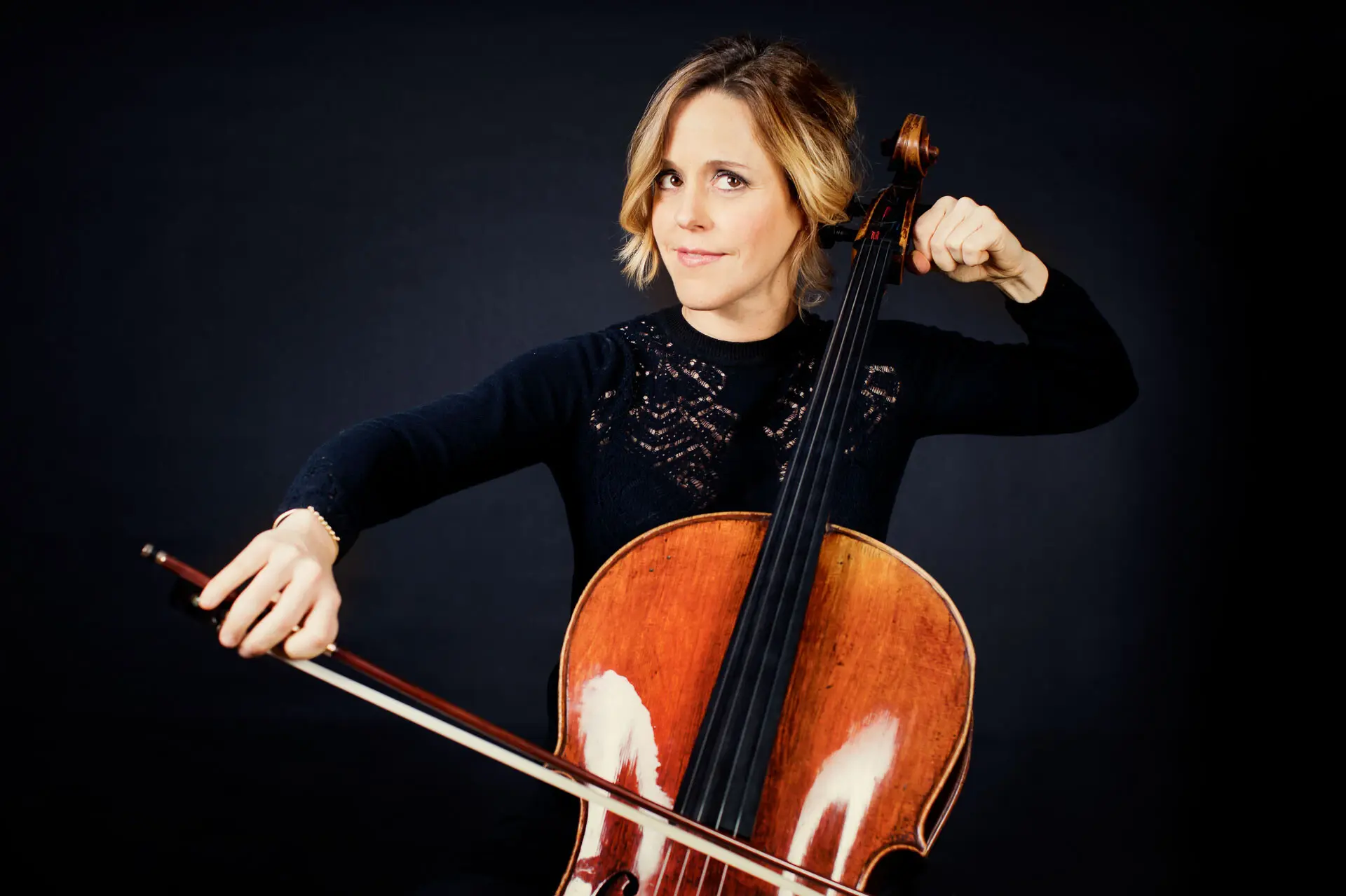 Vor einem dunklen Hintergrund sitzt eine Frau, die einen Cello spielt. 