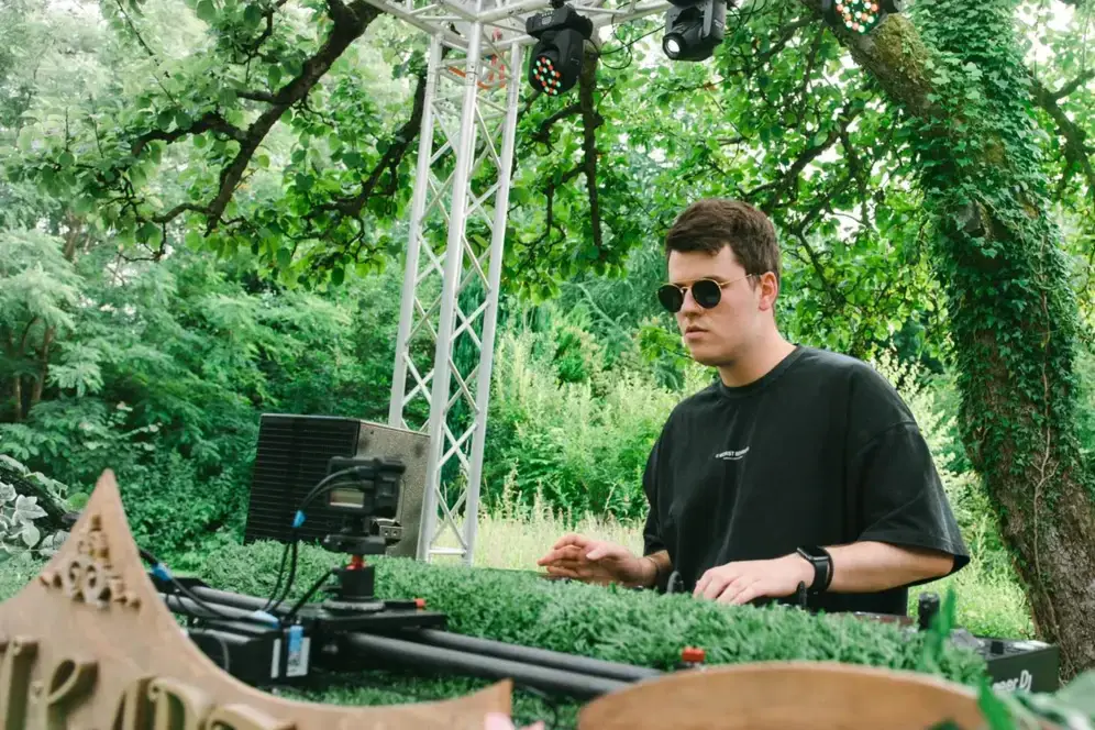 Ein Mann mit Sonnenbrille legt als DJ im Freien vor grünen Bäumen auf.