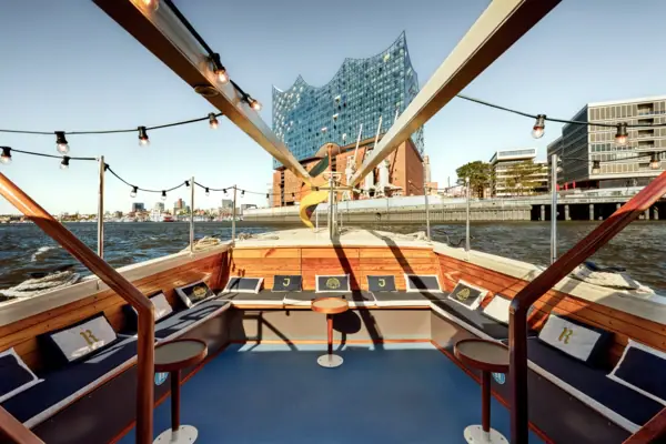 Der Außenbereich der Barkasse MS Jacob mit Sitzbänken und Kissen und der Elbphilharmonie im Hintergrund.