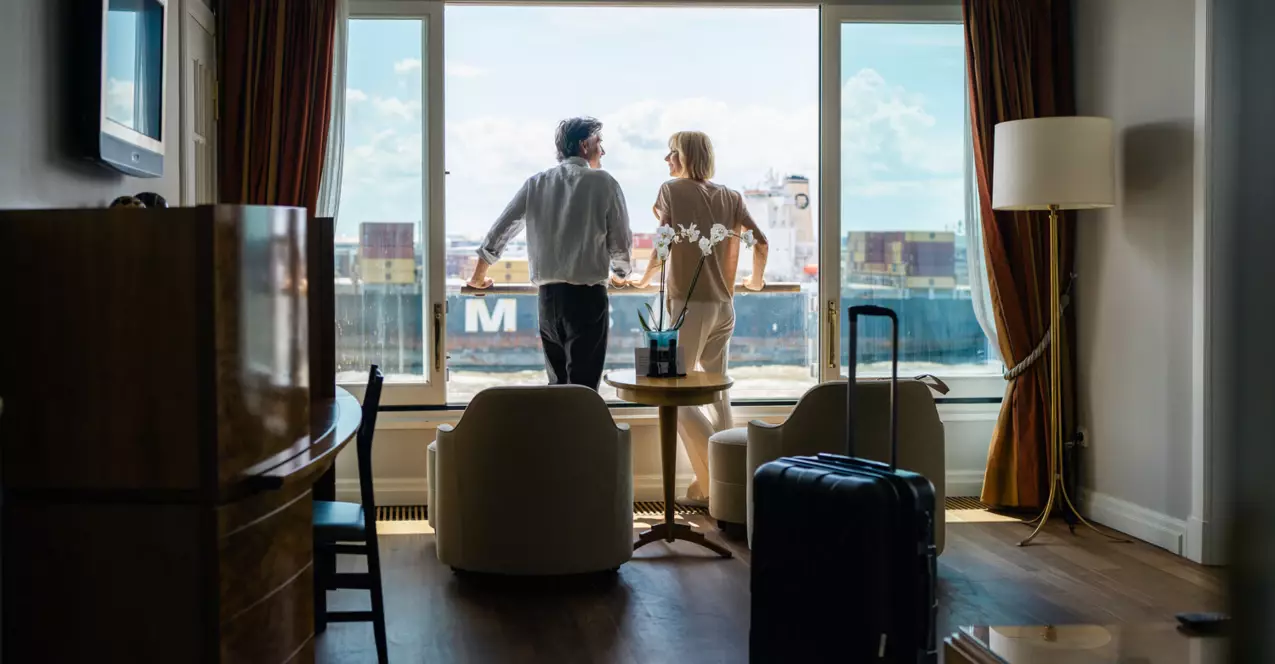 Ein Mann und eine Frau stehen an dem Fenster eines Hotelzimmer mit zwei Sesseln und kleinem Tisch sowie Schreibtisch und schauen auf die Elbe. Im Hintergrund befindet sich ein vorbeifahrendes Containerschiff und die Sonne scheint. 