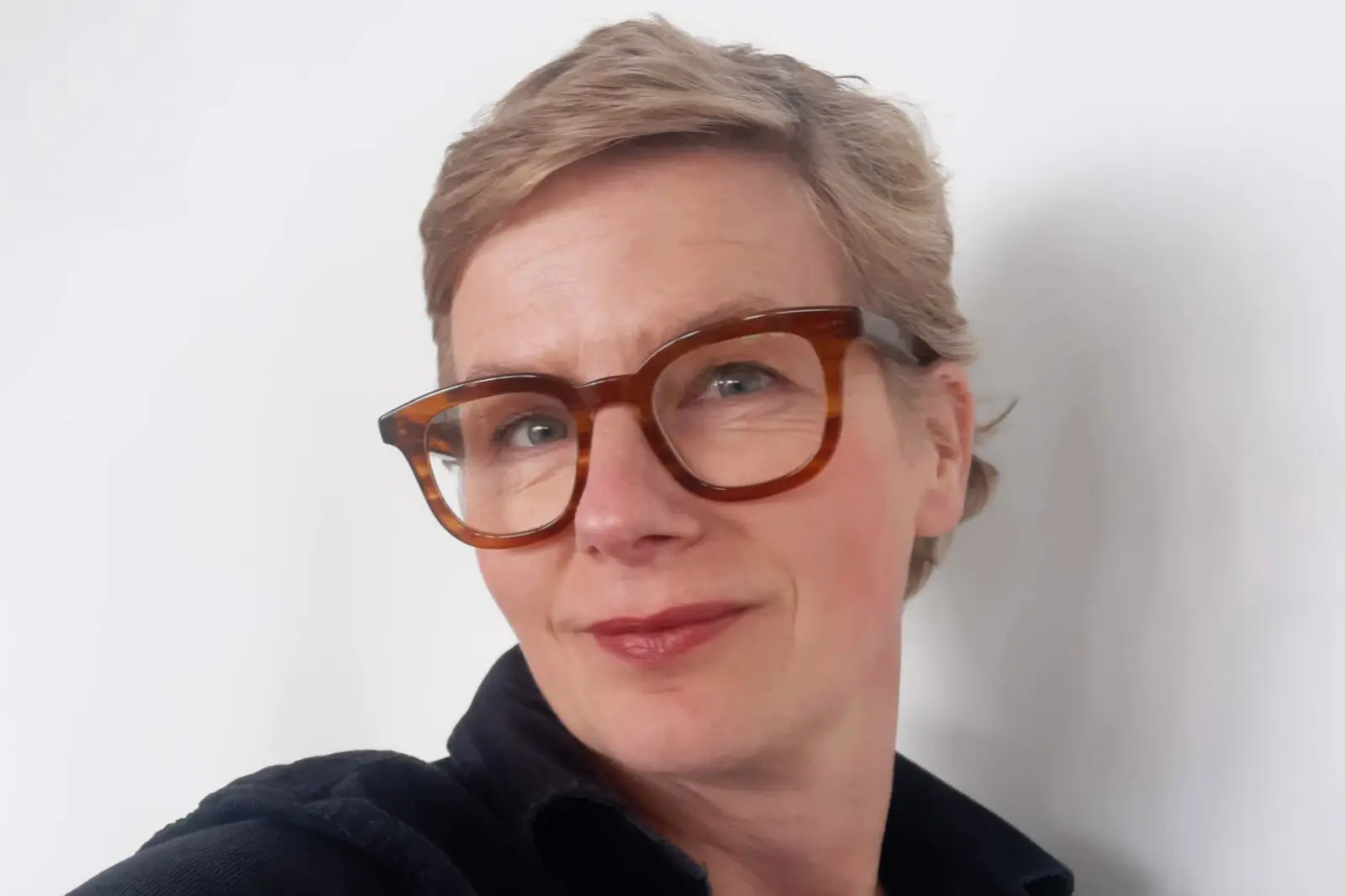 Eine Frau mit kurzen hellen Haaren und einer braunen Brille lächelt..