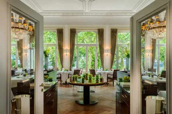 Blick in den Saal des Jacobs Restaurants mit Kronleuchtern, Tischen und Stühlen sowie dem Ausblick auf die Lindenterrasse durch die hohen Fenster.