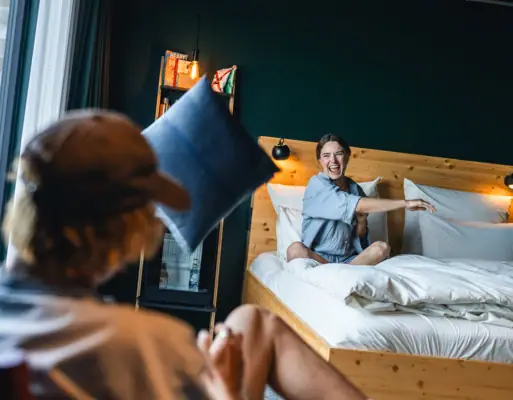 Zimmer im Urban Nature Hotel St. Peter-Ording Eine Frau sitzt auf einem Bett in einem Zimmer im Urban Nature Hotel St. Peter-Ording und wirft ein Kissen auf die Kamera zu.