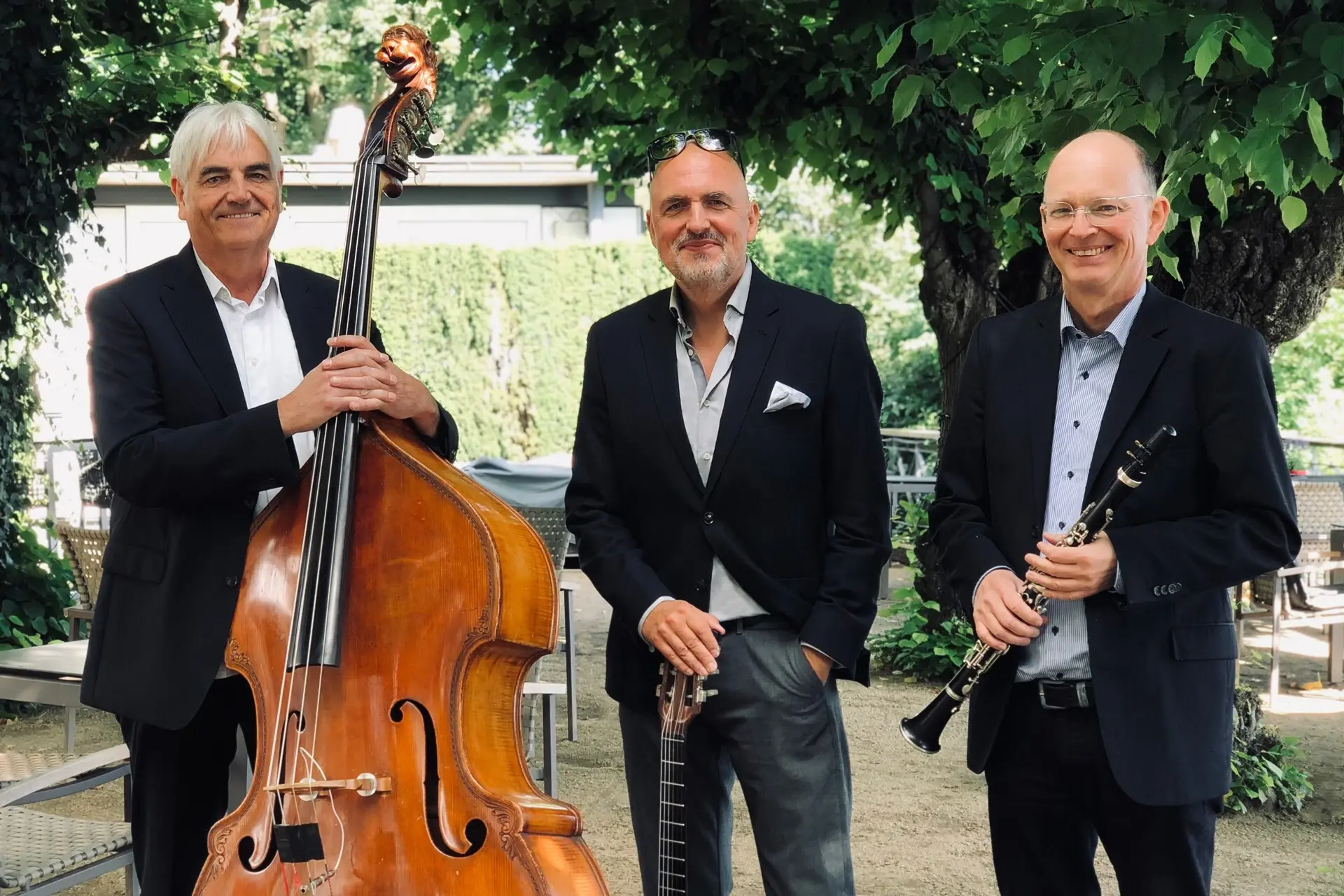 Drei ältere Männer halten jeweils ein Instrument, wie ein Cello, eine Gitarre und eine Oboe und lächeln in die Kamera.