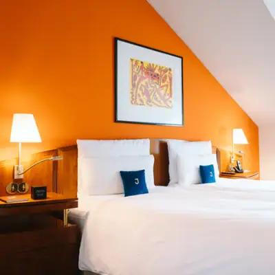 Bett in einem Zimmer des Hotels Louis C. Jacob vor einer orangenen Wand mit einem Pop-Art-Gemälde.