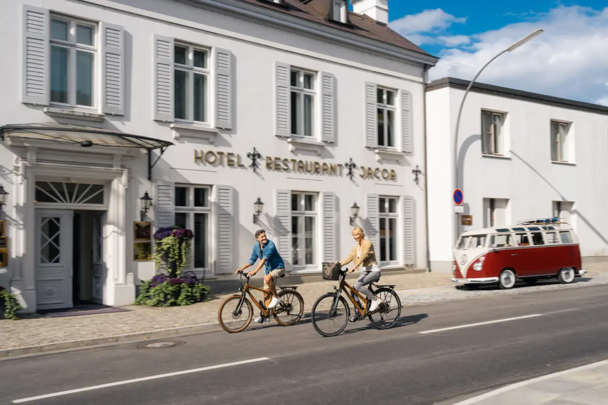 Ein Mann und eine Frau fahren Fahrrad auf der Straße vor dem Eingang zum Hotel Louis C. Jacob. 