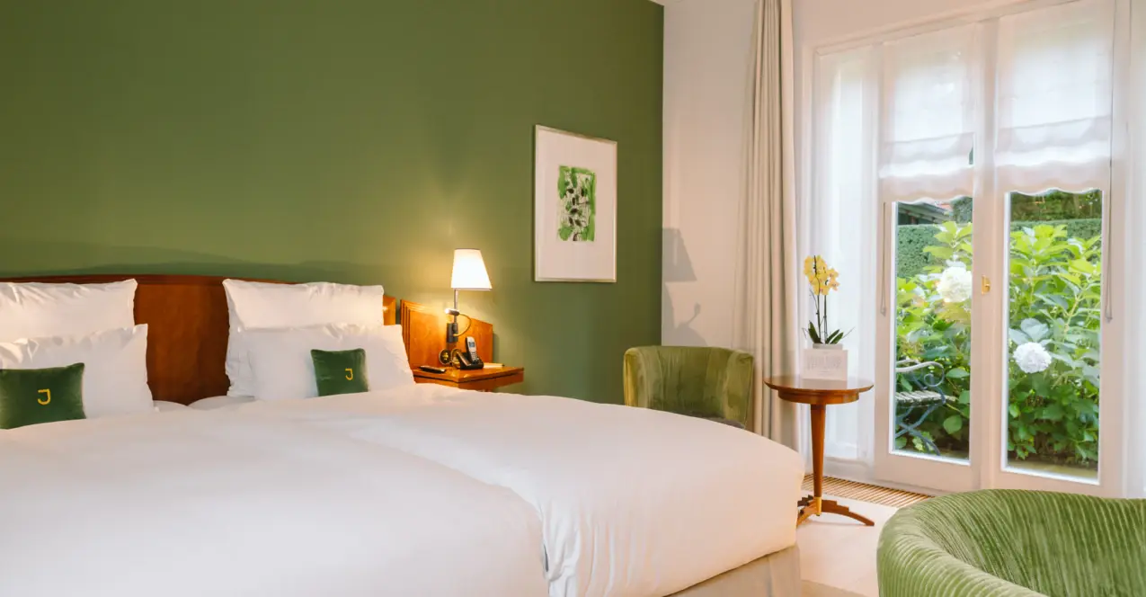 Stilvoll eingerichtetes Hotelzimmer mit grün akzentuierter Wand, gemütlichem Doppelbett, eleganten Sesseln und Blick ins Grüne durch bodentiefe Fenster.