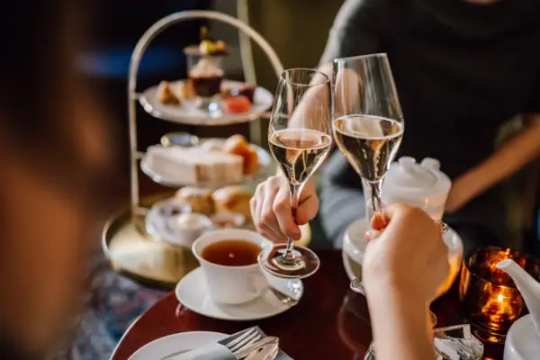 Afternoon Tea im Hotel Louis C. Jacob – auf dem Tisch steht eine Teetasse und zwei Personen stoßen mit Champagner an. Im Hintergrund sieht man eine Etagère mit Scones, Sandwiches und süßen Desserts.