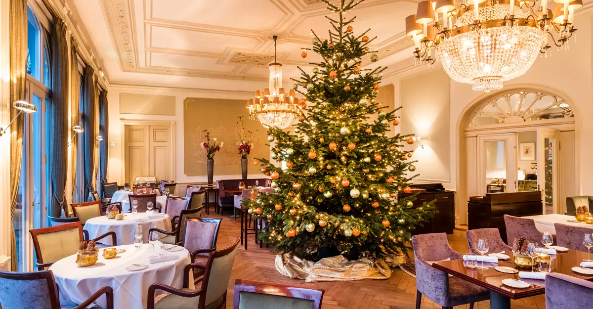 Ein Essenssaal mit hohen Wänden und Stuck. In der Mitte des Raumes steht ein geschmückter Weihnachtsbaum um den viele runden Tischen mit Stühlen stehen. Große Kronleuchter hängen von der Decke und füllen den Saal mit warmem Licht.