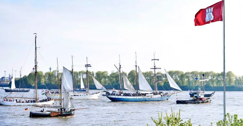 Eine Vielzahl von Schiffen beieinander in eine Richtung fahrend auf der Elbe. Im vorderen rechten teil des Bildes ist eine rote Hamburg Fahne gehisst und die Sonne scheint. 