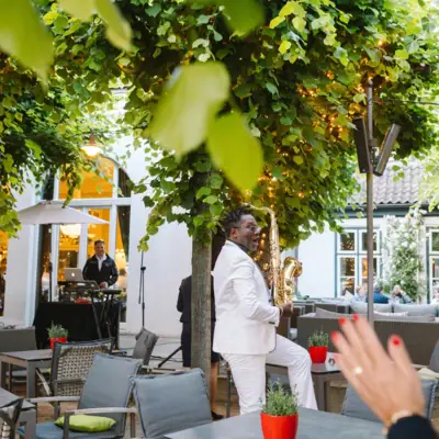 Ein Mann im weißen Anzug spielt Saxophon unter einem Baum.