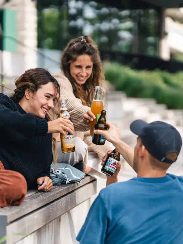 Terrasse Urban Nature St. Peter-Ording Eine Gruppe von Menschen sitzt auf einer Bank vor dem Urban Nature Hotel St. Peter-Ording und stoßt mit Bierflaschen an.